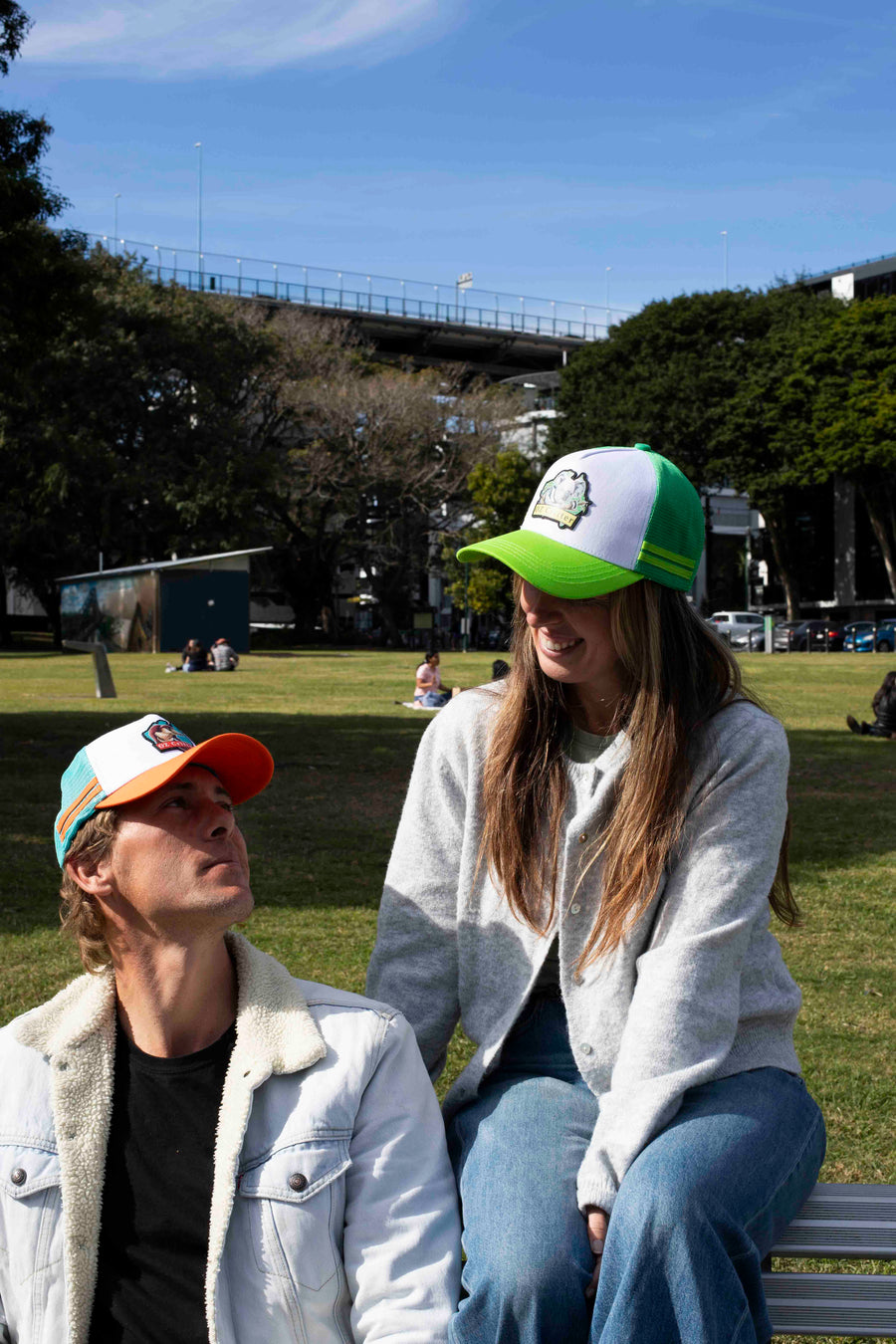 Two people sitting on a bench in a park with a bridge and trees in the background.