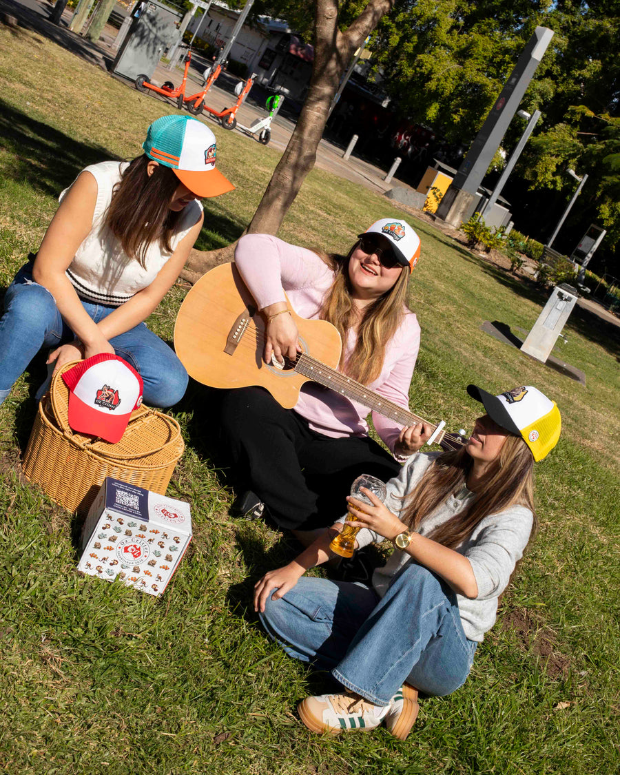 Three friends sitting on grass with a guitar, drinks, and a basket in a park setting.
