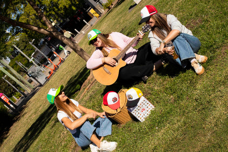 Three people sitting on grass playing guitar in a park wearing the OZ critter caps