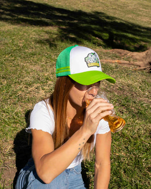 Person wearing a green and white Koala cap with OZ Critter logo, drinking from a glass outdoors on grass.