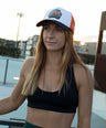 Woman wearing a black sports bra and green shorts with a fish cap, standing outdoors.