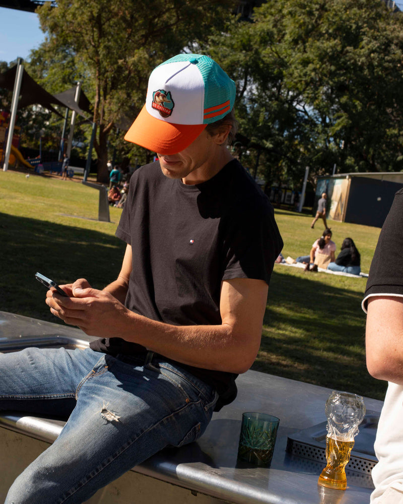 Man sitting outdoors wearing a platypus cap using a phone with a park setting in the background.