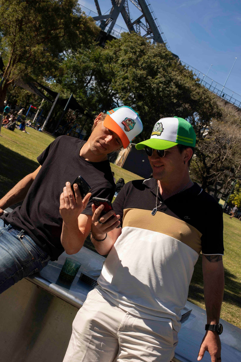 Two people sitting outdoors, wearing OZ Critter's cap one using a phone, with trees and a clear sky in the background.