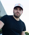 Man wearing a black t-shirt and white trucker cap with a logo outdoors