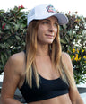 Woman wearing a black sports bra and platypus white cap with a logo, standing outdoors.