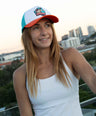 Woman wearing a colorful platypus cap and white tank top with a cityscape in the background.