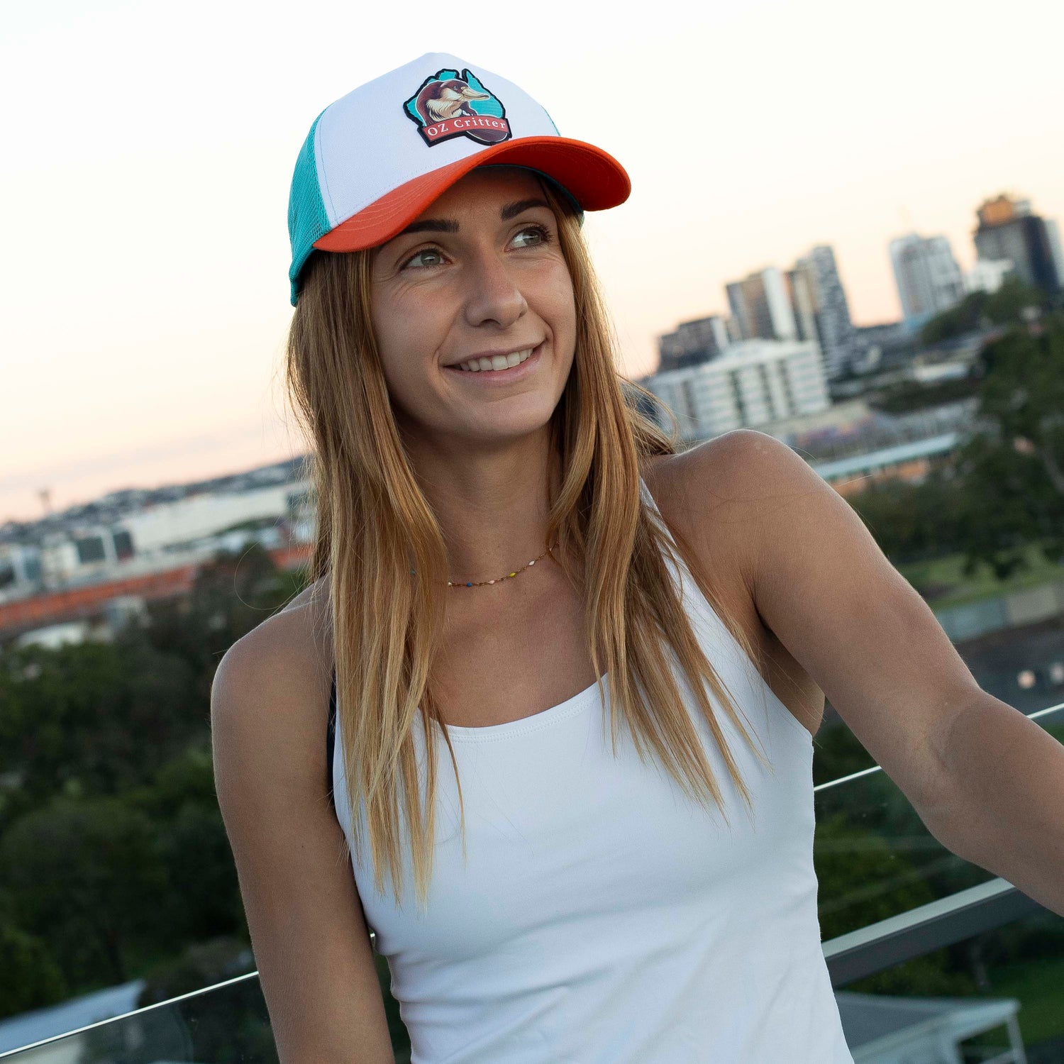 Woman wearing a colorful platypus cap and white tank top with a cityscape in the background.